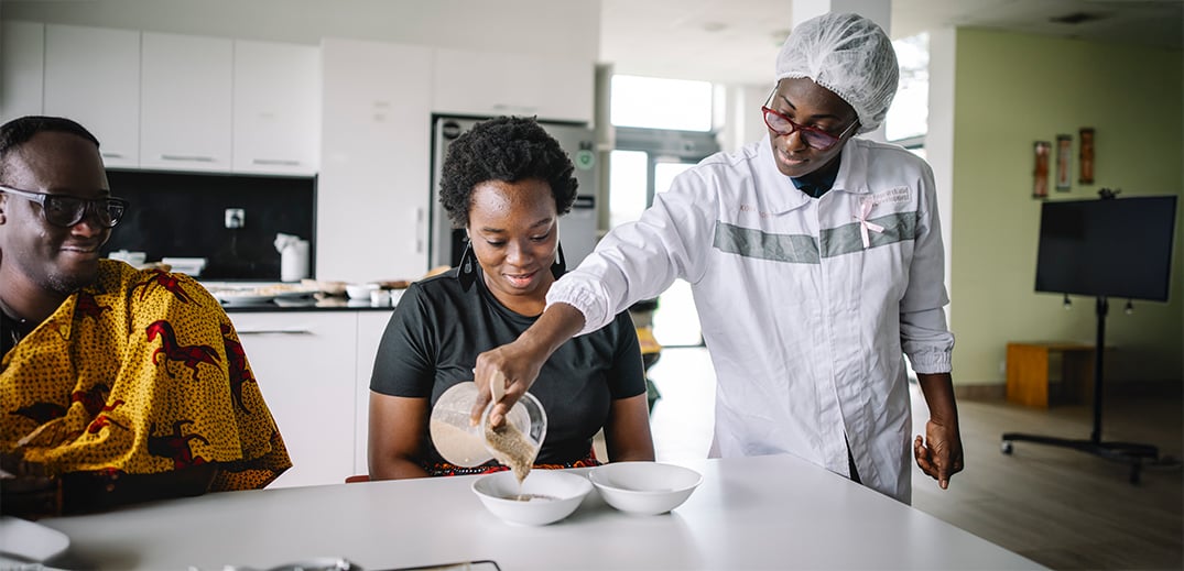 A person in a lab coat and hairnet pours liquid into a bowl for a seated woman, who smiles. Another person observes.