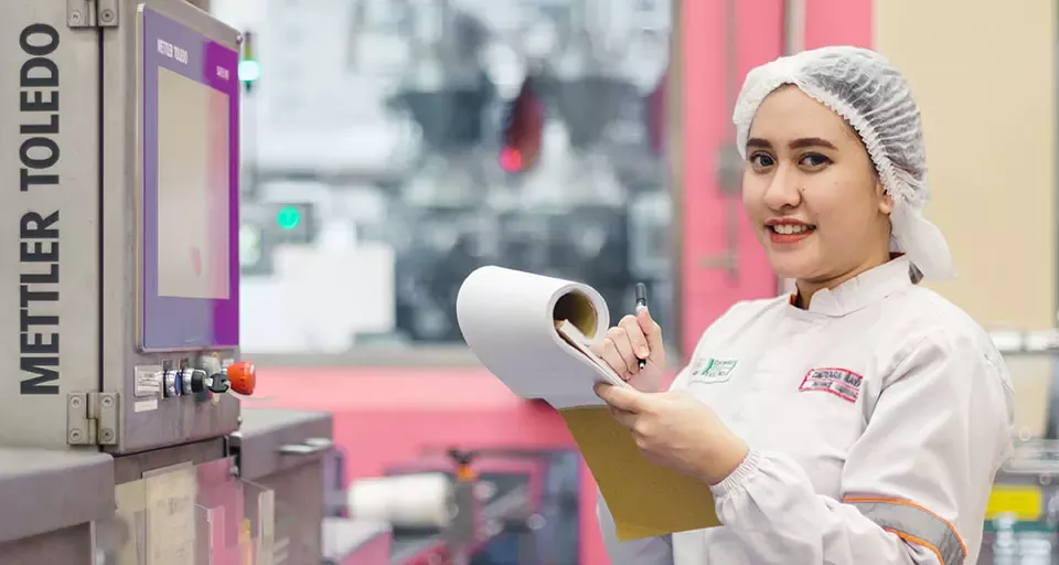 A worker in a lab coat and hairnet stands at a machine, holding a clipboard and pen, engaged in monitoring equipment.