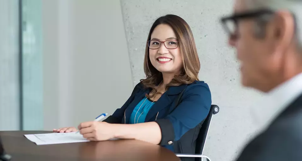 A woman sits at a desk holding a pen and paper, smiling, while another person in a suit is seated across from her
