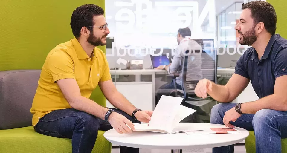 Two men sit on a green couch in an office, engaged in discussion.