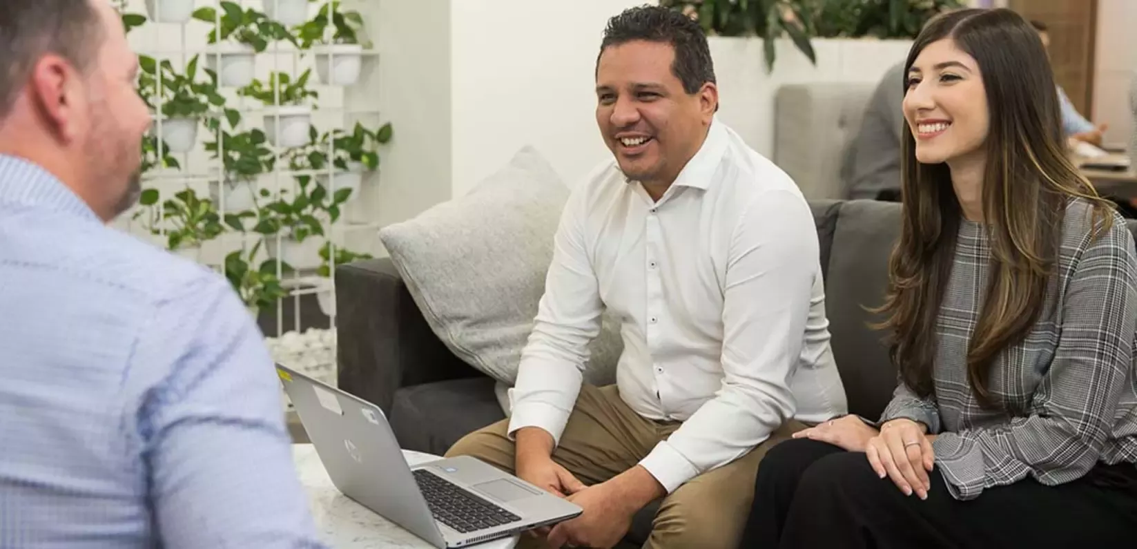 A group meeting taking place in a modern office setting, with a laptop on a table and indoor plants in the background.