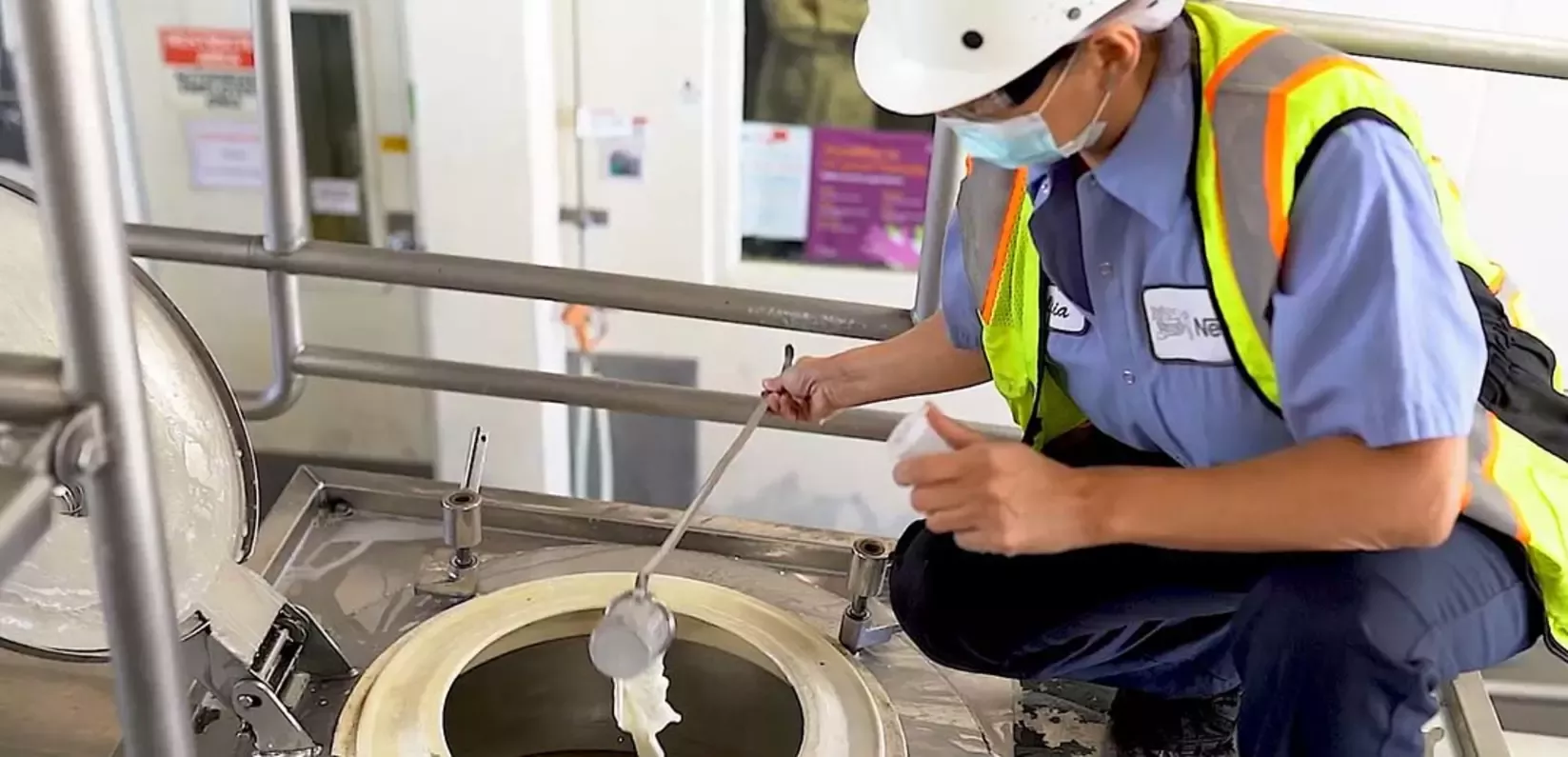 A worker in a safety vest inspects a large metal container, using a scoop to check the contents inside.