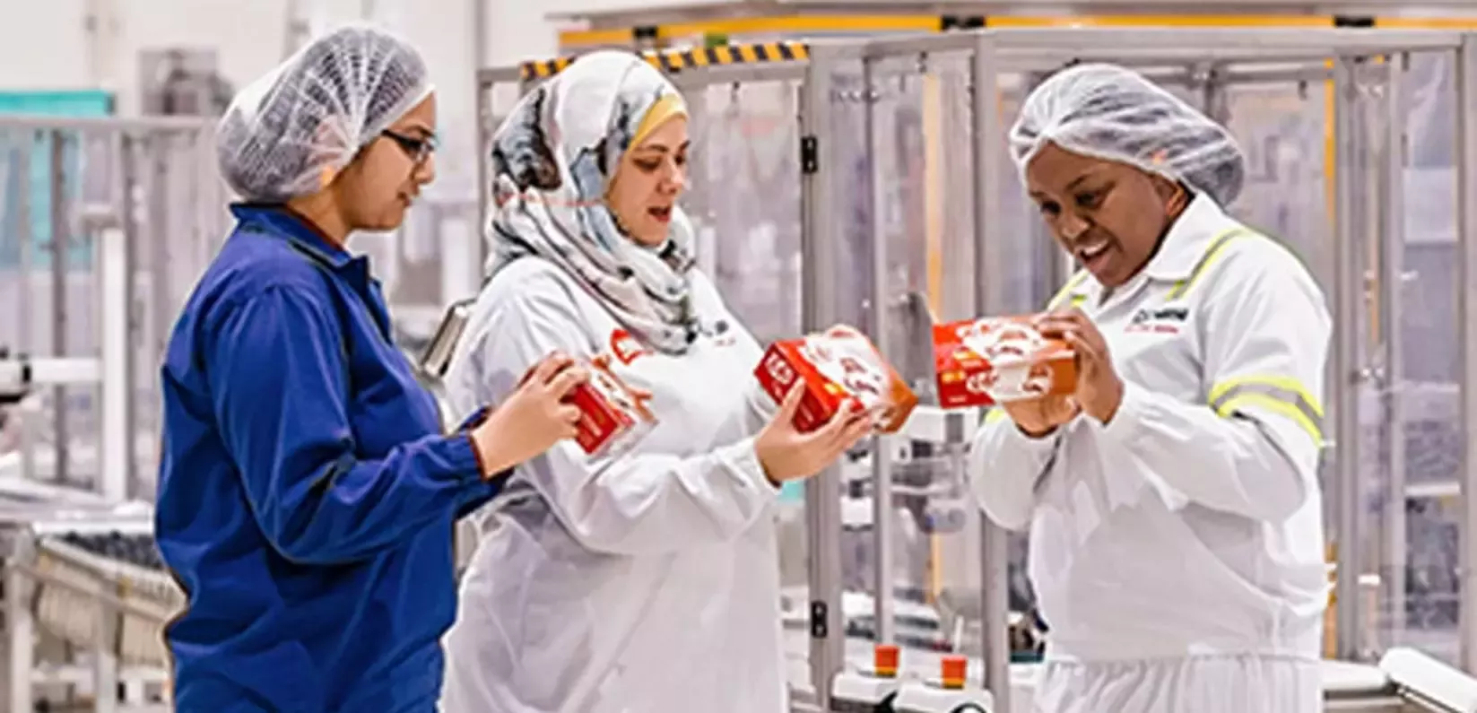Three factory workers in hairnets and uniforms are discussing products while holding red packaging in a production area.