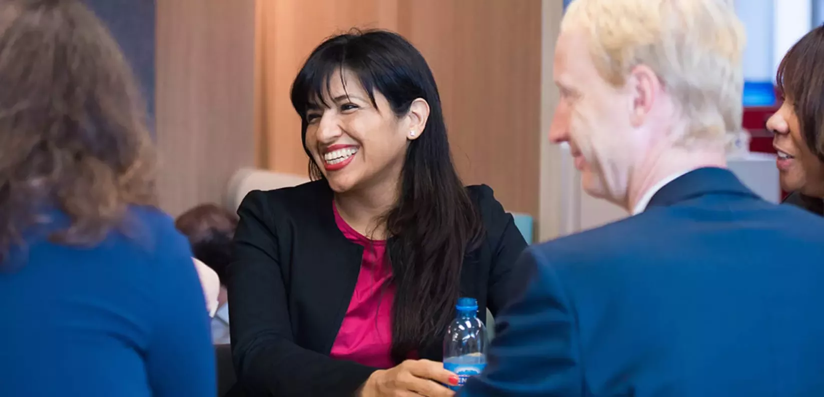 A woman in a black blazer and pink top smiles warmly during a meeting. She holds a water bottle.
