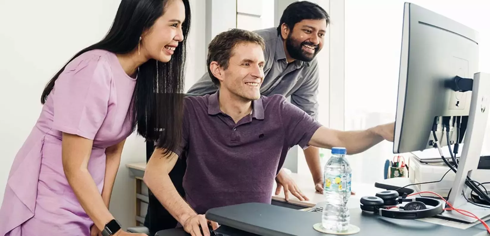 Three people are gathered around a computer monitor in an office, smiling while looking at the screen. 