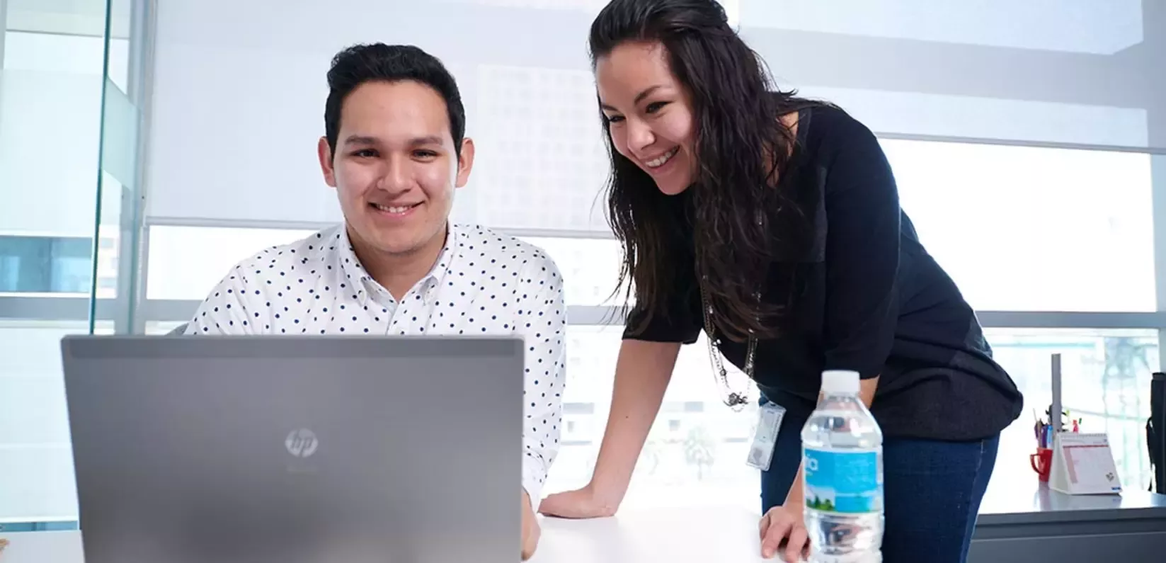 Two professionals collaborate at a desk with a laptop, while a water bottle and office supplies are visible in a bright, mode