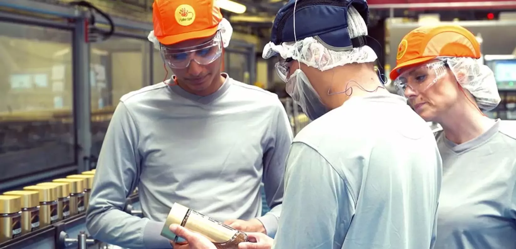 Three factory workers in protective gear examine a sealed product in a manufacturing setting with jars lined up in the backgr