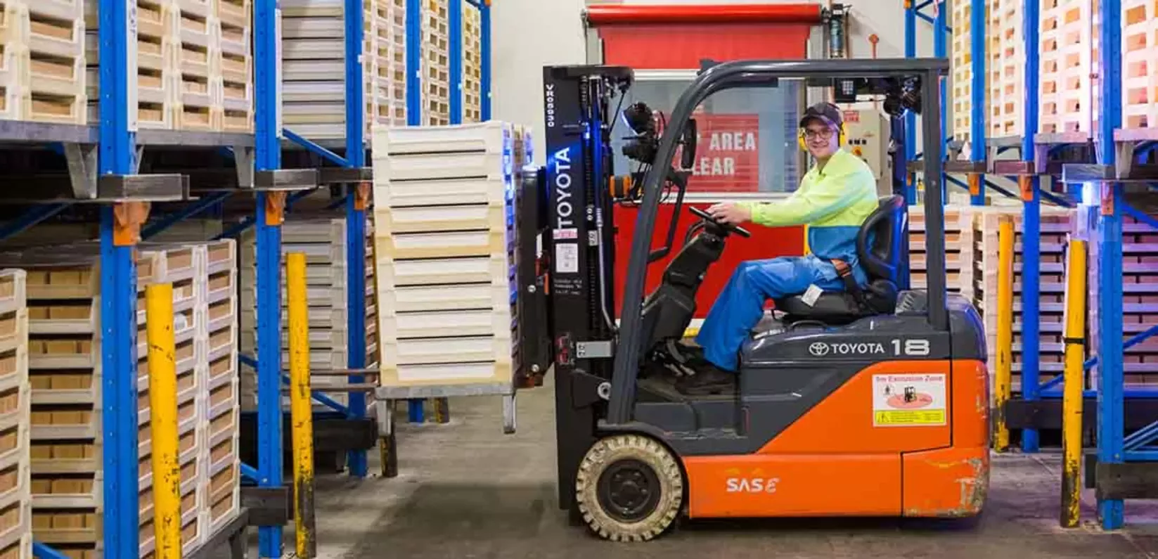 A forklift operator maneuvers a load of wooden pallets in a warehouse filled with shelves and safety markings.