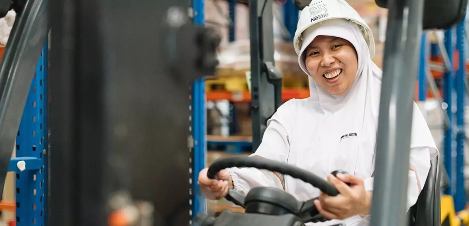 A warehouse worker in a white uniform drives a forklift, poised and focused, surrounded by shelves of stacked goods.