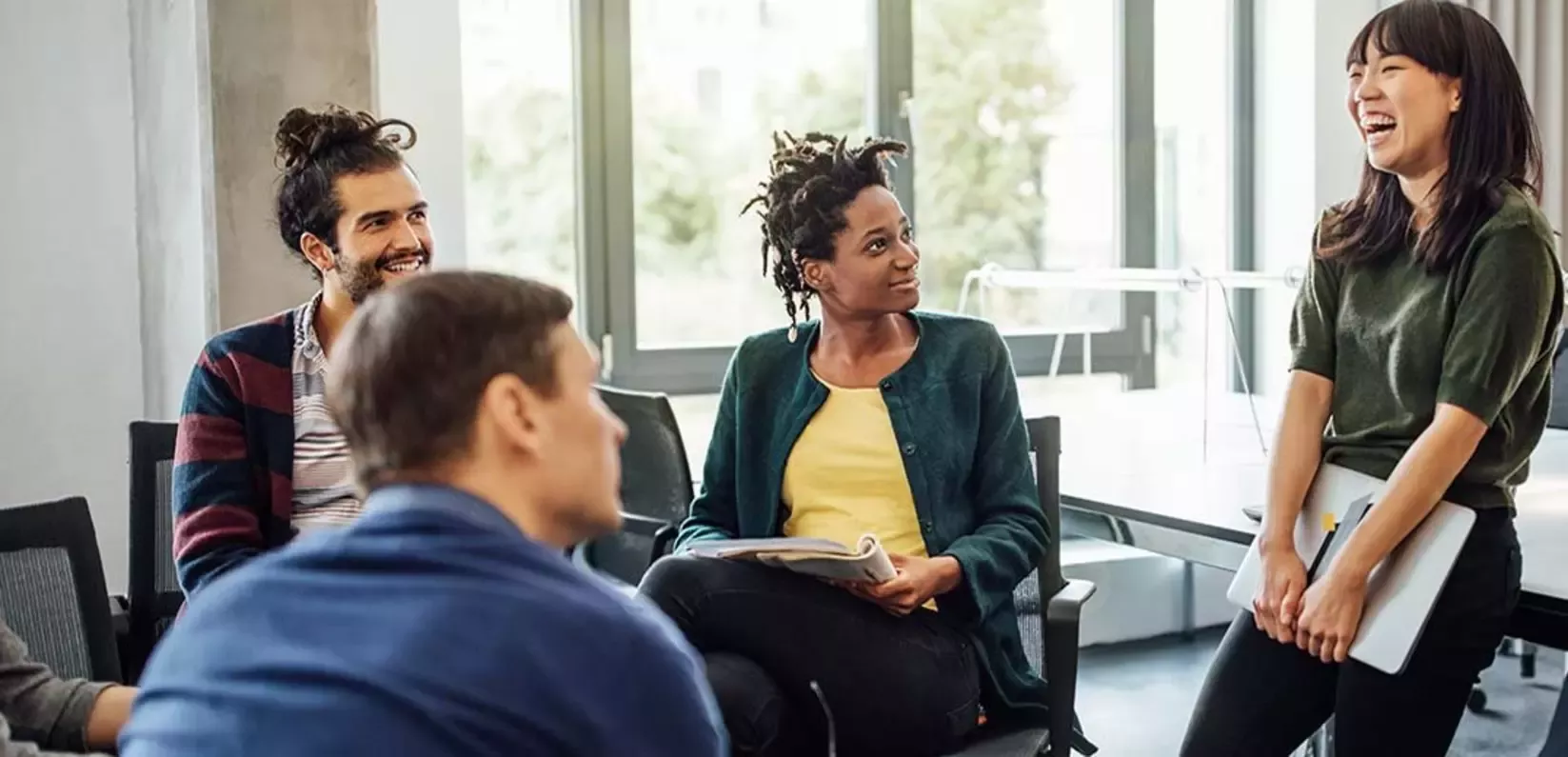 A group of diverse people engage in a meeting, seated in a bright, modern room with large windows 