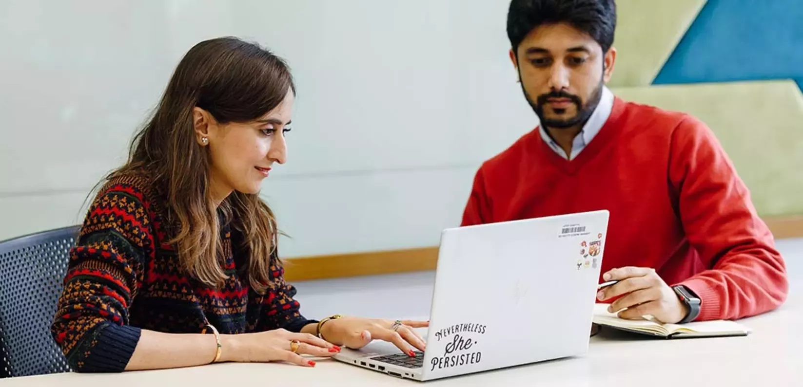 A woman with long hair in a patterned sweater collaborates with a man in a red sweater, both focused on a laptop 