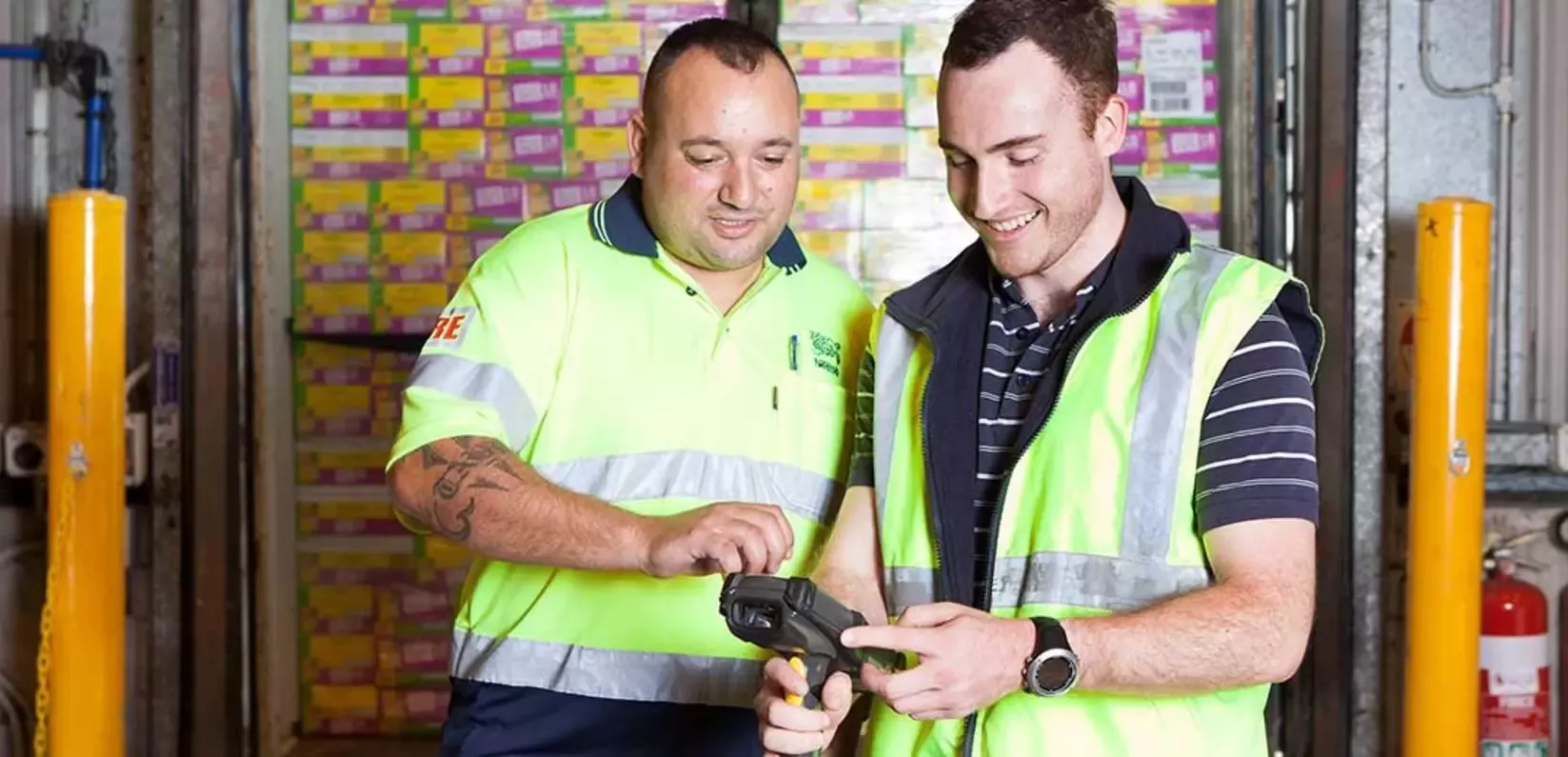 Two men wearing safety vests are focused on an appliance, likely reviewing information or instructions.