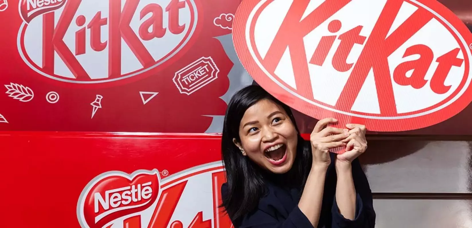 A woman holds a KitKat sign in front of a large box, smiling and promoting the chocolate brand.