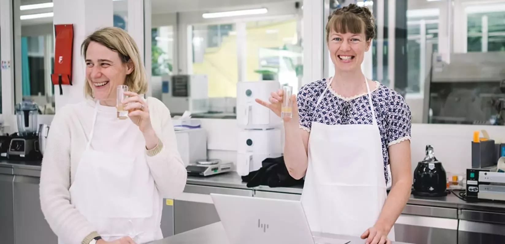 Two women in a bright room, wearing aprons, smiling while holding clear glasses. A laptop and lab equipment are visible on th