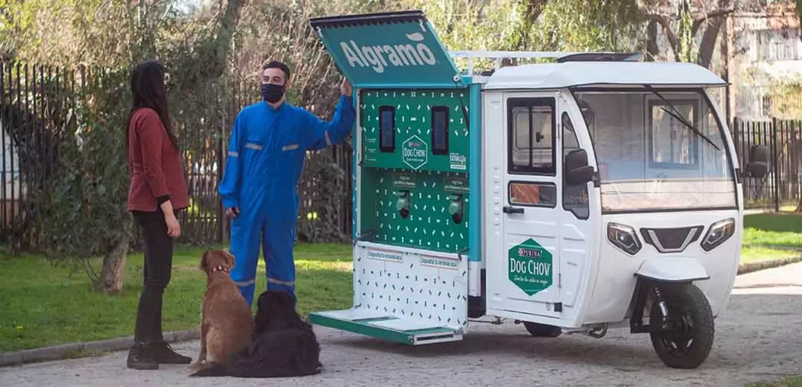 A person in a blue jumpsuit and mask stands by a white vending tricycle labeled "Algramo," speaking to a woman 